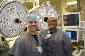 Two CRNAs posing in an operating room with large surgical lights behind them