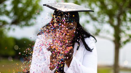 woman wearing white coat and graduation cap. She is blowing confetti toward the camera