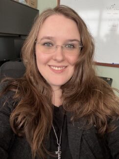 Head-and-shoulders portrait of a woman in her mid-thirties with long reddish brown hair, looking at the camera in soft lighting.