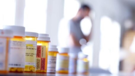 A large group of prescription medication bottles sit on a table as a man in the background stands with his hand on his head. The image is photographed with a very shallow depth of field with the focus being on the pill bottles in the foreground.