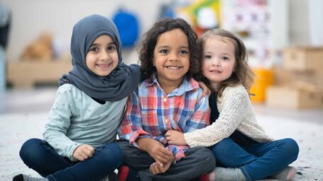Two girls and a boy are indoors in their preschool classroom. The multi-ethnic group is smiling at the camera while sitting on the carpet and embracing. One girl is Muslim and she is wearing a hijab and the other girl is Caucasian.