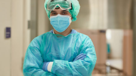 Partial front view of male surgical attendant in early 20s wearing protective clothing, eyewear, mask, bouffant cap, and looking at camera with arms crossed.