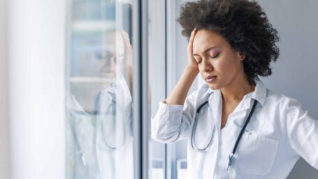 Tired female doctor near window in hospital. Female doctor with head pain standing near window. It's been a physically and emotionally challenging day