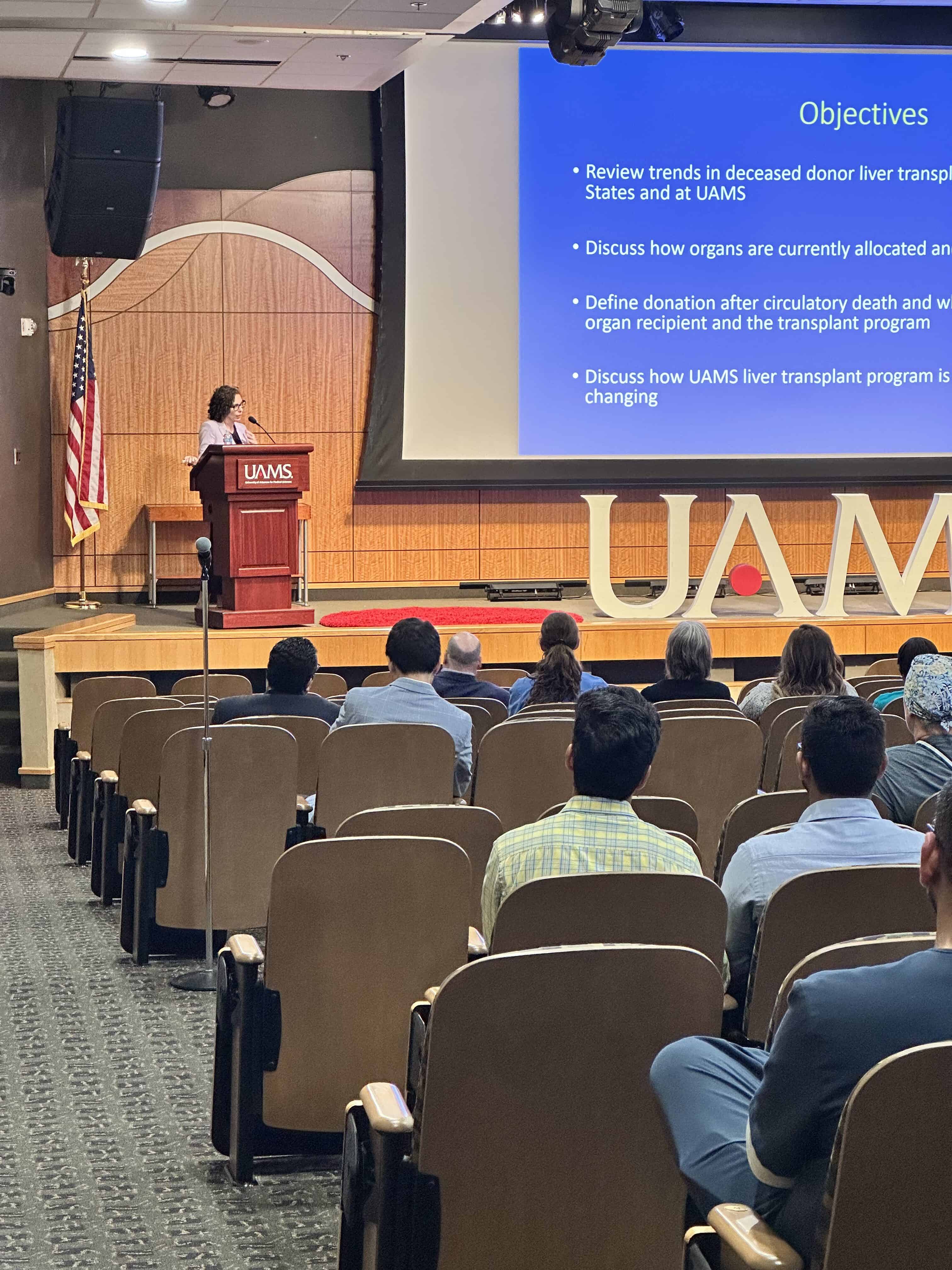 Speaker stands at a podium next to a large video screen with presentation notes. People sit in the auditorium in front of her listening.
