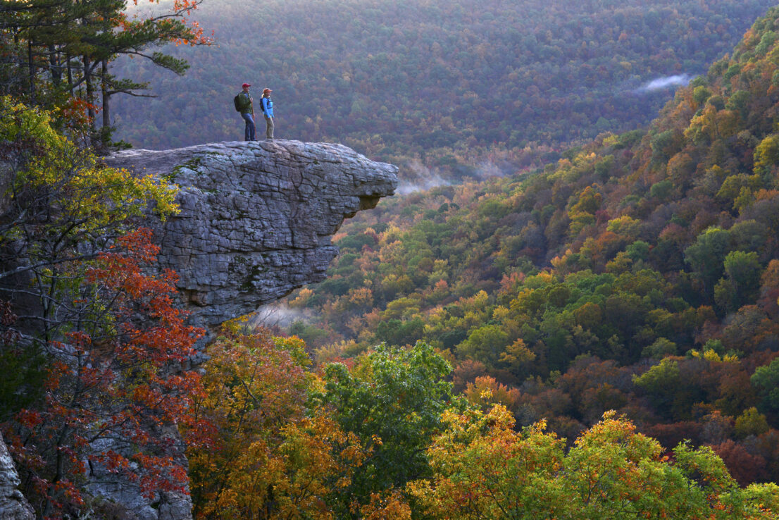 Shot of hikers on Hawksbill Crag during the fall. Colorful trees are in the distance.