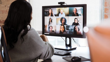 A group of diverse business colleagues participate in a virtual staff meeting during the COVID-19 pandemic. An African American businesswoman participates in the virtual event from her office.