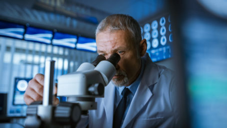 Senior Medical Research Scientist Looking under the Microscope in the Laboratory. Neurologist Solving Puzzles of the Mind and Brain. In the Laboratory with Multiple Screens Showing MRI / CT Brain Scan Images.