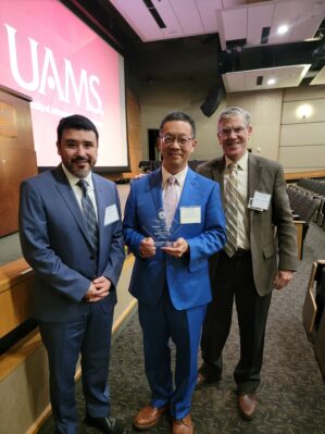 Department chair and faculty member posing with Dr. Lai. Dr. Lai is holding a plaque.