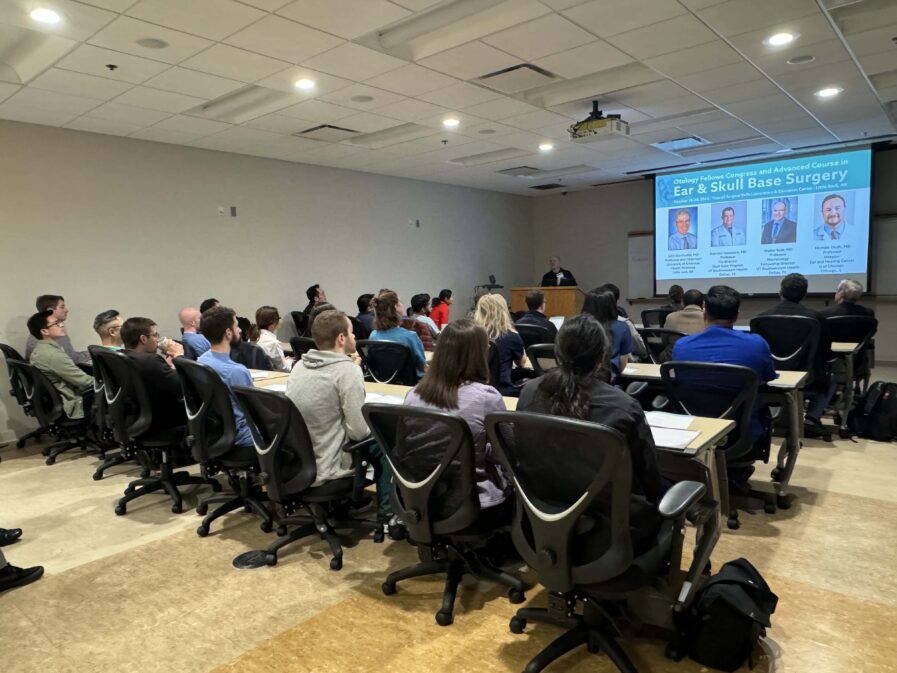 Group of people in a lecture room listening to a talk. The speaker is standing behind a lectern and beside a large video screen