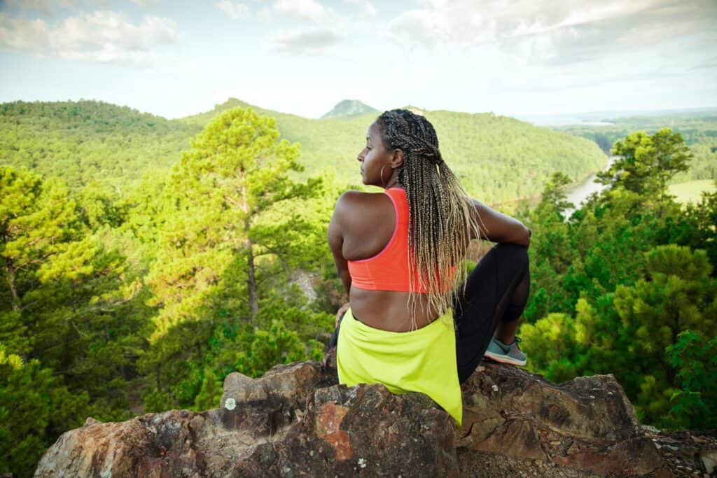 A Black female hiker rests on a rock outcropping taking in the view from the side of Pinnacle Mountain