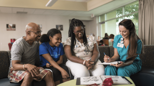 Family of 2 parents and a young girl sit on a couch and speak with a nurse in green scrubs taking notes on a clipboard