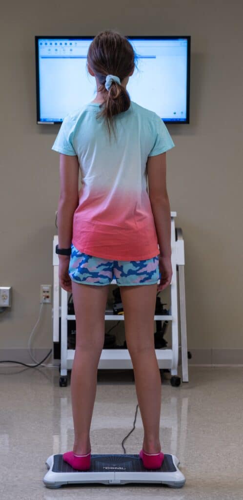 back of a school age girl standing on a balance plate facing a screen