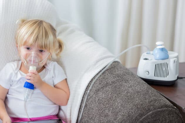 Caucasian preschool age girl on sofa receives breathing treatment. Her blond hair is in pigtails.