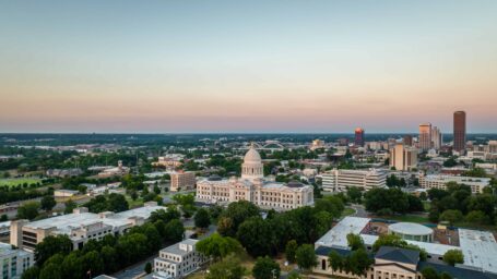 A view of downtown Little Rock at sunset from a drone. The Arkansas State Capitol Building stands in the foreground, the gold point of its dome standing out against the Broadway Bridge's white arches.