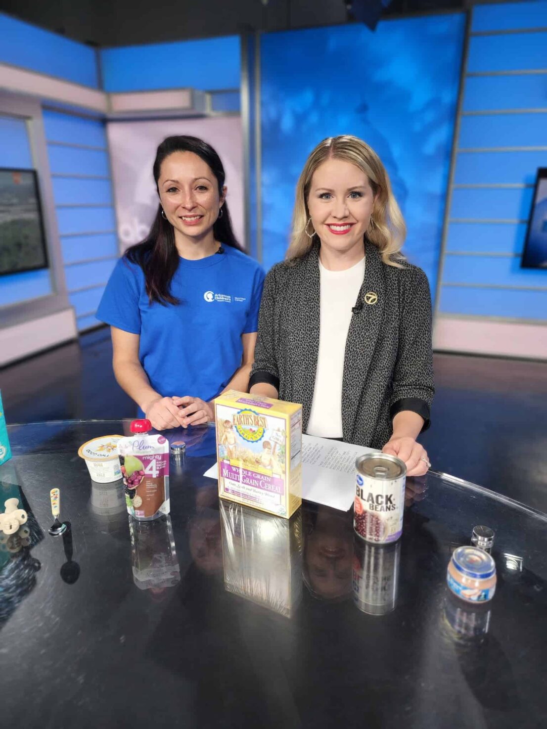 an ACNC dietitian poses next to a news correspondent smiling in front of formula milks and first food box mixes on set for the morning news