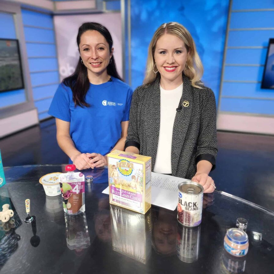 an ACNC dietitian poses next to a news correspondent smiling in front of formula milks and first food box mixes on set for the morning news