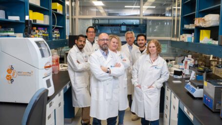 7 men and women in white lab coats standing arms crossed in a lab