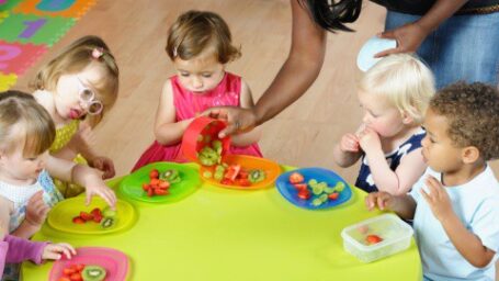 A portrait of a care giver serving fruit to a group of toddler/ children in a nursery setting.