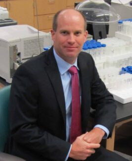 Jeffery H. Moran, Ph.D. Dr. Moran in his lab wearing a black suit - Blue shirt with a red tie. Sitting in front of a collection beakers and lab cylinders