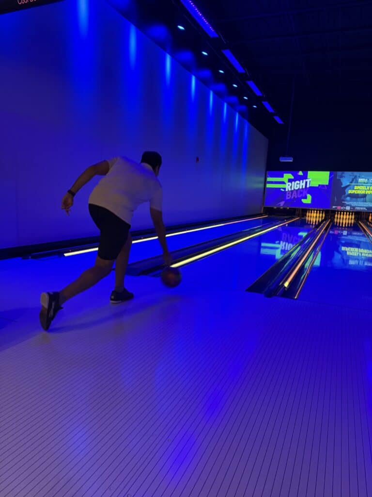 Bowler releases a ball down a neon-blue bowling lane, with glowing pins and colorful screens ahead; the dark venue feels energetic and modern.