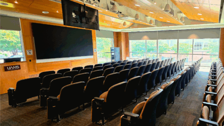 Modern lecture hall with rows of black chairs, large windows, a projector screen, ceiling-mounted monitors, and wood paneling. Natural light fills the room.