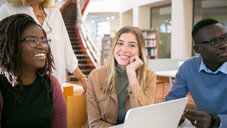 A diverse group of university students sitting at a table with a laptop, smiling while listening to an instructor in a modern library.