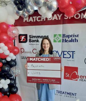 Caitlin Caperton holding up a poster showing that she residency matched in Psychiatry at UAMS. She is standing in front of a festive banner surrounded by balloons
