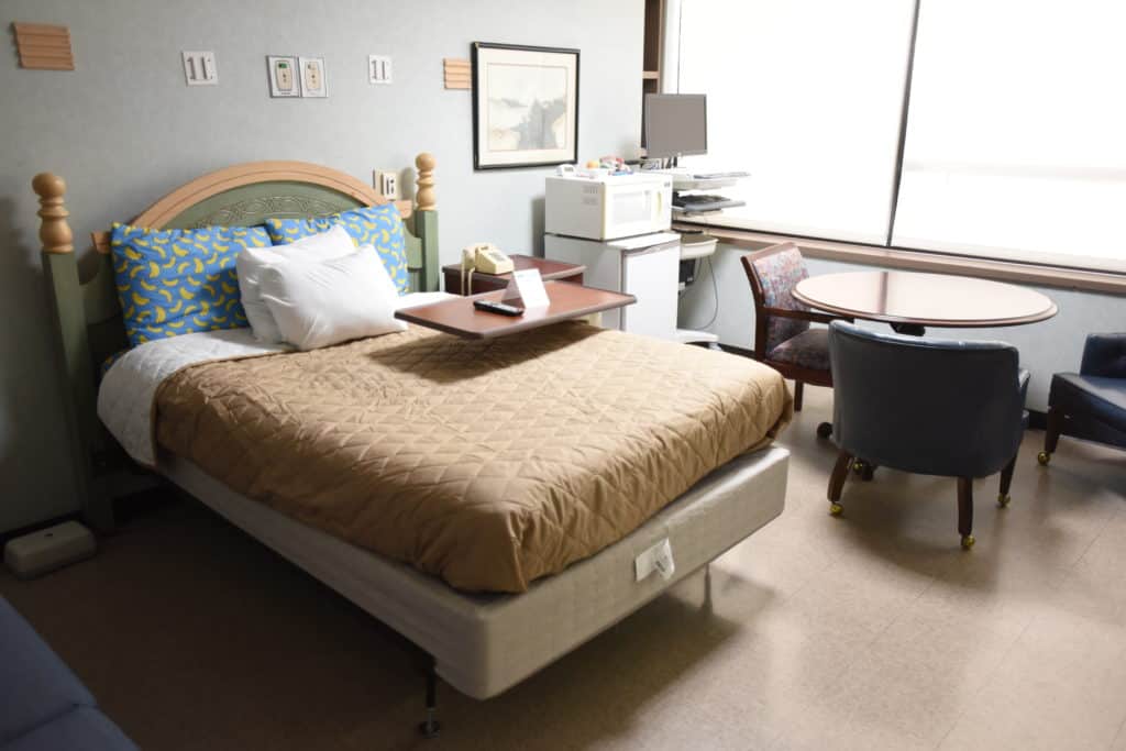 Interior of a call room at the Baptist Health Rehabilitation Institute. Picture includes a bed, small table with chairs, dorm-sized fridge with microwave, and a computer station.