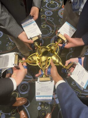Close up of students' hands holding trophies and their conference name tags. The photo is taken from above, and the students are holding the trophies together.
