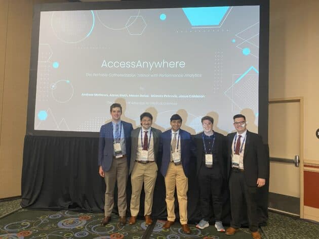 Group photo of students standing in front of a large video screen that reads "AcessAnywhere" The Portable Catheterization Trainer with Performance Analytics"