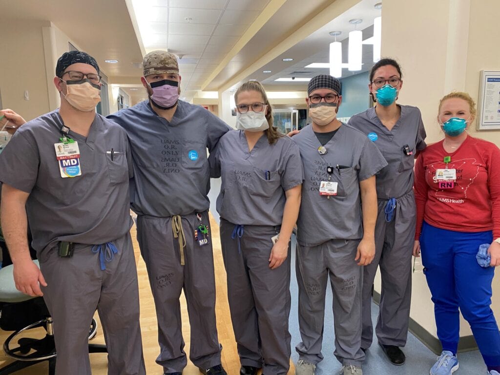 Group of faculty and fellows posing in a hospital hallway. They are wearing scrubs and masks.