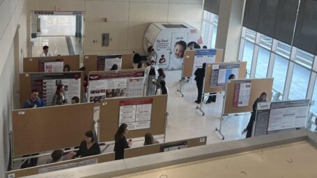 Overhead shot of the room, showing all the research posters mounted to rolling bulletin boards. A student is standing by each one, explaining their research to onlookers.