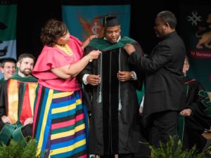 Two people assisting student put on regalia hood over gown