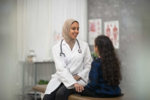 A female Muslim doctor sits up beside her Hispanic young female patient as she talks with her about the appointment. She is wearing a white lab coat, a Hijab, and has a stethoscope around her neck. The young girl is sitting cross legged in front of the doctor as she listens attentively.