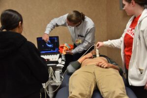 Faculty member holding a heart model and pointing to an image on a screen while a student uses an ultrasound to image a volunteer's heart