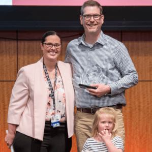 Mary Katherine Kimbrough, Chris Cathcart holding award, and Chris's toddler daughter on stage