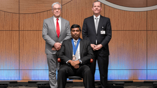 Dr. Shashank Kraleti seated in commemorative chair on stage, flanked by Dr. Steven Webber and Dr. Cam Patterson