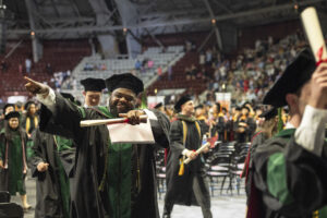 Graduate, wearing regalia, points as he walks in a line with fellow students