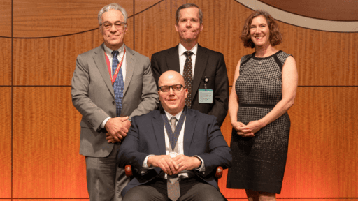 Dr. Ashley Acheson seated in ceremonial chair on stage, flanked by Drs. Steven Webber, Cam Patterson and Laura Dunn