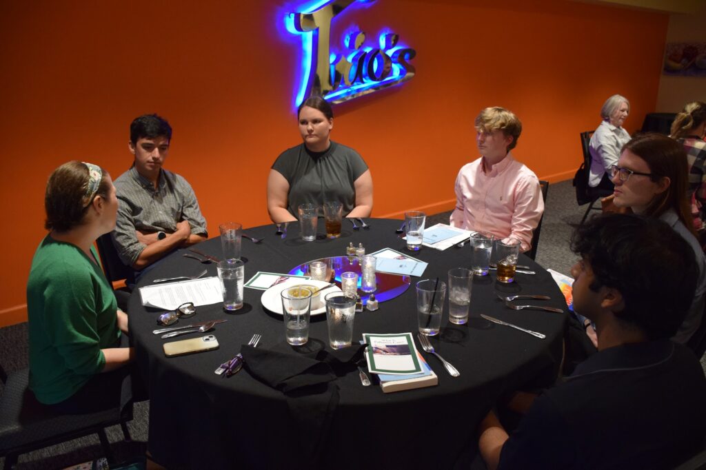 
Medical students gather around a round table at Taco's restaurant during a palliative care discussion event, with educational materials visible on the table.