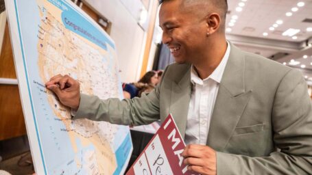 Man in a suit smiling and placing a pin on a large USA map, holding a sign about matching in Pediatrics. The mood is celebratory and joyful.